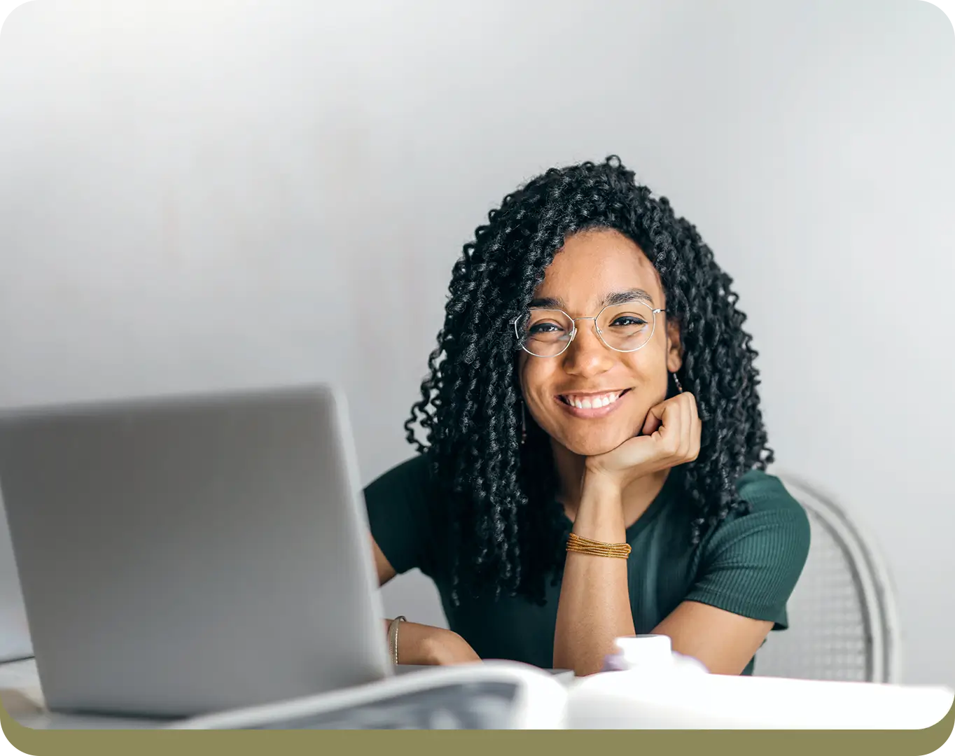 A person with curly hair and glasses sits at a desk, smiling, with a laptop in front of them.