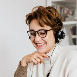A person with short hair and glasses smiles while wearing headphones, sitting in the PI Online Training Office, a well-lit room with shelves in the background.